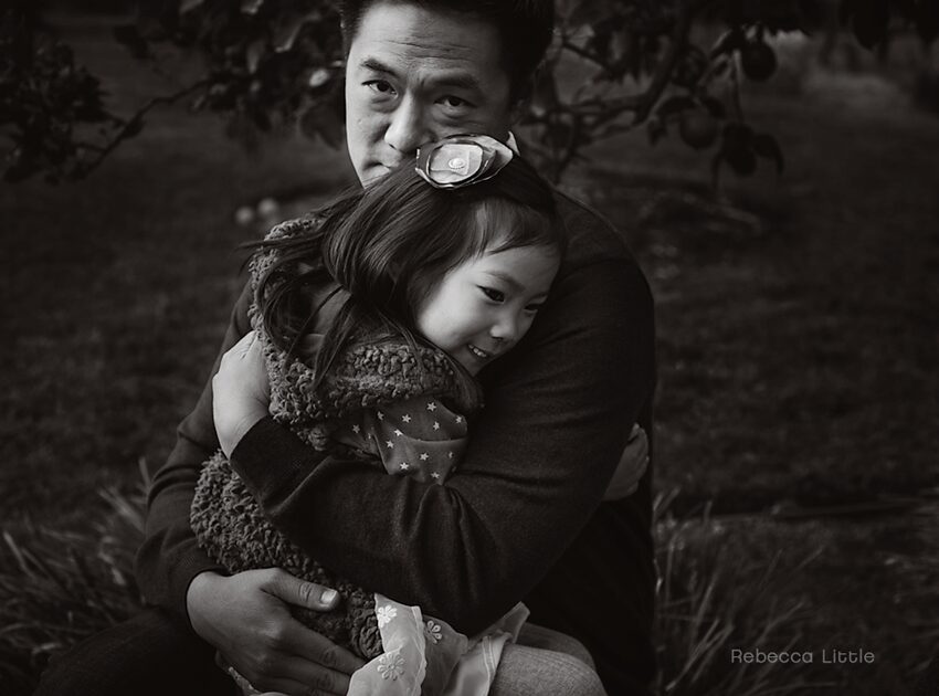 Black and white holiday mini sessions sneak peek. This is an image of a father and daughter. The young girl is sitting on her father's lap and he is hugging her. Photos for Christmas cards. Photos for Hanukkah cards. Image by Rebecca Little Photography Pasadena, CA.