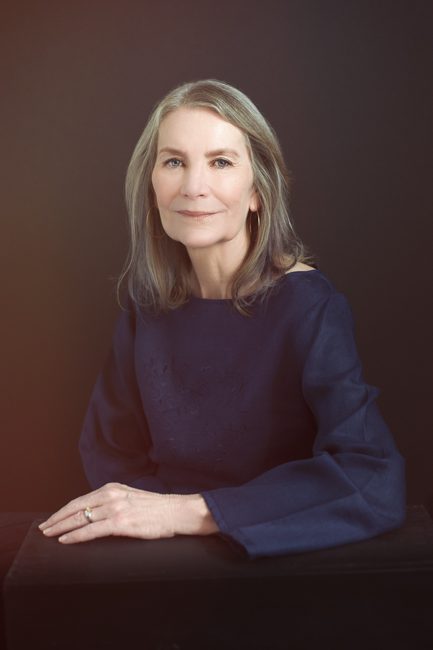 This headshot is of an Executive Director of a foundation. She is transitioning into consultant work and wanted an image that was conservative and elegant and professional. She's wearing a deep blue linen blouse that features a boatneck and long, wide sleeves. he hand and arm ar reasting on a black table. Her body is slightly angles away from the camera. Her face is looking towards the camera and she has a slight smile. her gray hair reached her shoulders and softly swept back from her face. The backdrop is black hand-painted canvas, the same kind used in Vanity Fair magazine style portraits. The overall feel of the image is understated elegance. This headshot was created by Rebecca Little Photography Pasadena, CA.