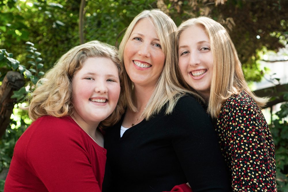Mother and her two daughters at a mini session in Pasadena. These three blondes are so well coordinated. Mom is wearing a black sweater, the younger sister is wearing a red sweater, and the older sister is wearing a red, black, and white patterned dress. They have their arms around each other and are standing very close. Image created by Rebeca Little Photography Pasadena CA.