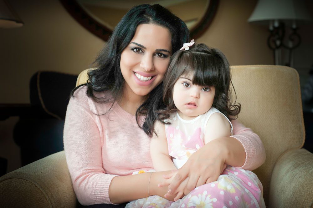 This is an adorable portrait of a young mom and her toddler daughter. They are sitting in a soft, gold suede armchair. The girl is sitting in mom's lap. The mom is wearing a light pink sweater with a scoop neck and three-quarter sleeves. Her daughter is wearing a sleeveless pink dress decorated with white daisies with yellow centers. The mom has long black hair and dark eyes. She has a beautiful smile as she looks at the camera. Her daughter is looking at the camera but not smiling. This family photo was taken at the Langham Hotel in Pasadena, CA by Rebecca Little Photography.