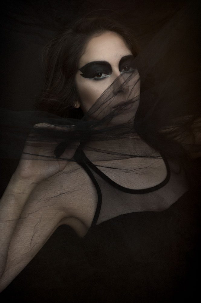 This is an image of a young woman peeking through black sheer tulle. We created a dark angel portrait for her personal use. She's wearing a black sleeveless dress with a tulle bodice. Black tulle is blowing across her face and she has heavy black eyeliner that covers her entire lid, up to her eyebrows. The has on tiny silver earrings. Her hair is long and black and blown out straight. This alternative portrait was created by Rebecca Little Photography in Pasadena, CA.