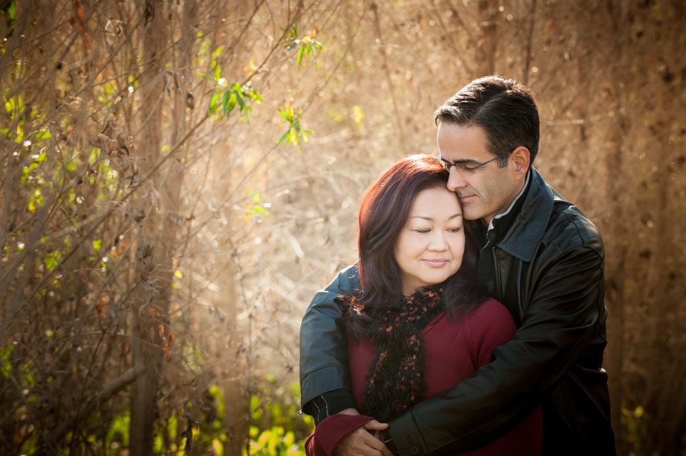 A man and woman photographed outdoors by Rebecca Little Photography. This is from a Mini Session Altadena session. The man is behind the woman - his wife - and has his arms around her. His face is nuzzling her hair. He's wearing jeans and a black leather jacket. She is wearing a red sweater and patterned scarf. She's looking away from the camera a slightly back at him. They seem lost in thought. A stand of trees is behind them.