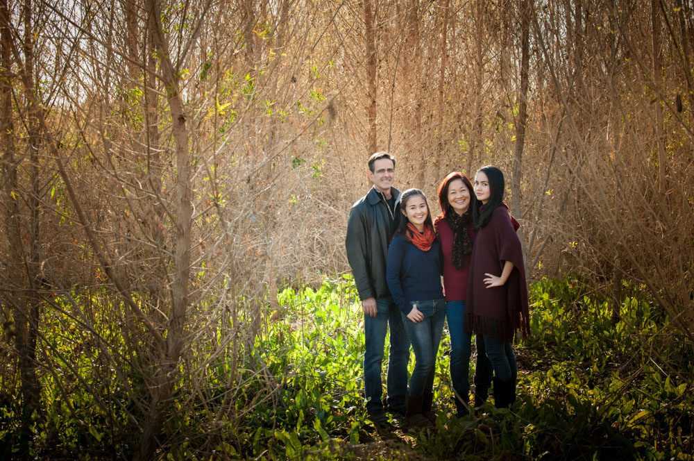 This beautiful family photo was created at one of my Mini Sessions Altadena. There is a family of four standing outside among a stand of trees. There is a man, woman, and their two daughters. They are dressed in jeans and jewel-toned sweaters. This is a wide shot, so they only fill one quarter of the frame and they are on the right side of the frame. The rest of the frame is filled with tall slender trees and  green shrubbery. Family photo created by Rebecca Little Photography Pasadena, CA.