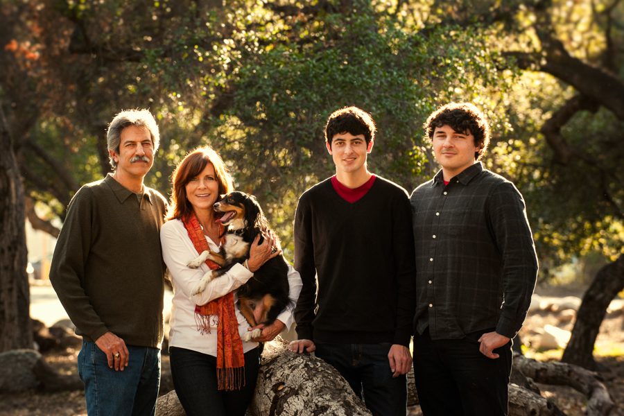 This is an image of a family of five with their dog. This is a Holiday Mini Session for a La Canada family. They are outside with trees behind them. The setting sun is behind them and filtering through the trees. The sun creates a golden halo around their heads as it glimmers on their hair. They are dressed in casual clothes in neutral colors of brown, gray, and black. The woman is wearing a white sweater with a red scarf and she is holding the small dog. Image created by Rebecca Little Photography.