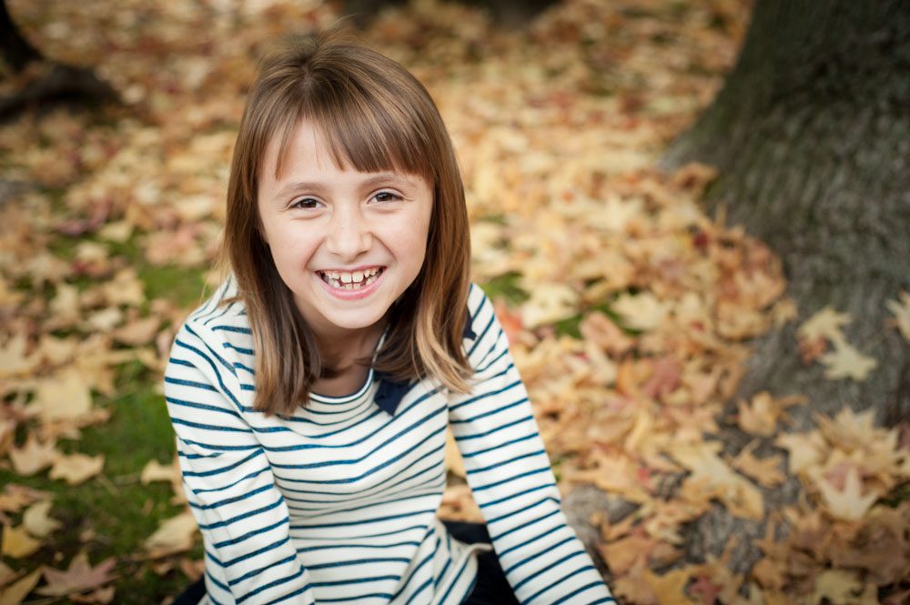This image is from a mini sessoin in San Marino. This young girl is sitting on the ground in a pile of brown leaves. She's looking up and smiling at the camera. She's wearing a blue and white horizontal striped sweater. He smooth brown hair curves at her shoulders. Photo created by Rebecca Little Photograph Pasadena, CA.