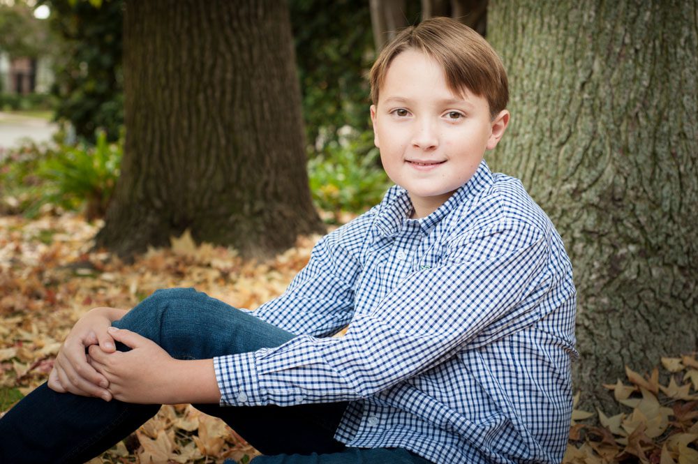Young San Marino boy sits on the goround near the base of a tree. He's wearing a blue and white checked shirt and dark blue jeans. There ground is ocvered with brown fall leaves. He is smiling at the camera. Photo taken by Rebecca Little Photography Pasadena CA