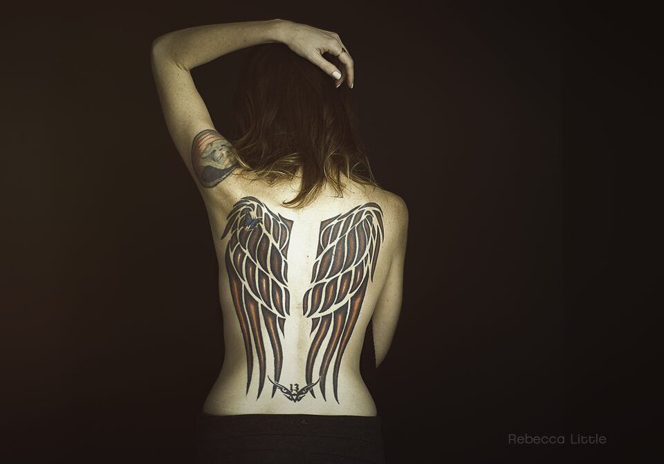 Octavia loves her tattooed back, just one of her many tattoos. This image shows her back with a full back tattoo of wings. She is only wearing black pants. We can see the curve of her hips. Her left arm is raised up and her arm is curved over her head. The backdrop is black and the only thing visible is her back. The light is highlighting her skin and throwing everything else into darkness. Creative headshots and cool branding for artists in Los Angeles. 