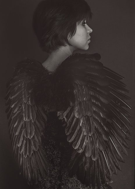 Sam is wearing black feathered wings. Her back is towards the camera and she is looking off to her right. The backdrop is black. Her wings are highlighted by the light shining on them. It's a moody, dark, serious, contrasty photograph.This is a black and white image was created by Rebecca Little Photography Pasadena, CA.