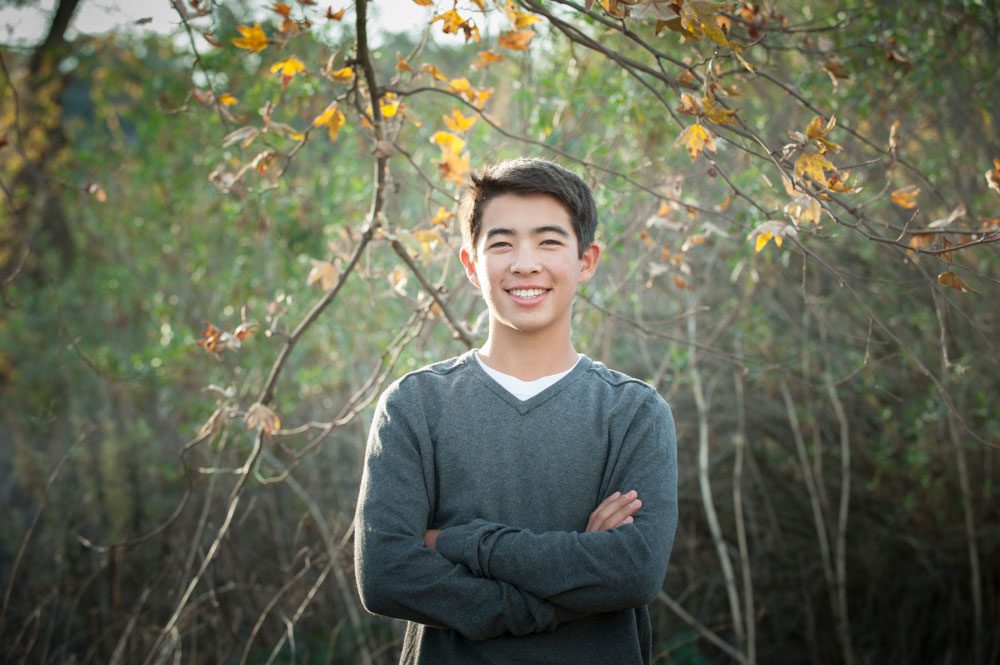 This is a photos of a teenaged boy during a family session in La Canada. He's standing in front of trees in a wooded area. There are a few green and gold leaves. He has on a pale blue gray sweater, his arms are crossed, and he's smiling at the camera. Image created by Rebecca Little Photography Pasadena, CA.