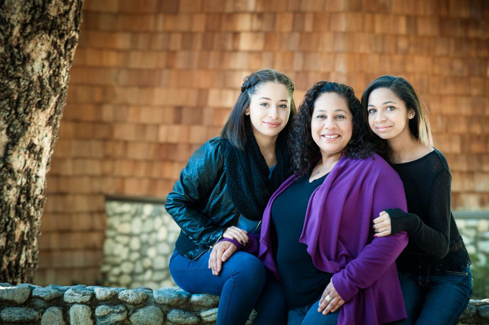 This family photo was taken during a Mini Session in Sierra Madre. We went to Little Santa Anita Canyon in Sierra Madre to create this image. It's of a mom and her two school-aged daughters. There is a house with wood shingles and a low stone wall behind them. They are sitting on a stone wall in the foreground. The mom has a bright purple sweater and black blouse, and the daughter are wearing jeans and black tops. They are connected by Mom's hand resting on her daughter's leg and the other daughter behind her hugging her arm. They are smiling at the camera. Image taken in Sierra Madre by Rebecca Little Photography.