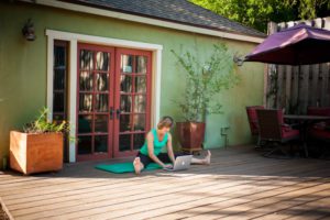 Pilates instructor Kelly sits on her outside deck and works on her computer. She sits on her wooden deck with her studio behind her. The studio is painted green on the outside and there are several potted plants. The large French doors are painted brick red. Branding photos taken by Rebecca Little Photography at Kelly's Altadena studio.