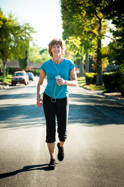 This is an image of a woman in her 40's running on a neighborhood street. She's wearing black yoga pants and a bright blue tshirt. She has an iPod in her hand and earbuds in her ears. It's a sunny day and she casts a long shadow in the street. It is about 5 pm in southern California where this shot was taken. She is smiling at the camera and it looks like we caught her mid-workout. This is a full-length body photo and illustrates outfits for dating photos for an active person. Image created in Pasadena, CA by Rebecca Little Photography.