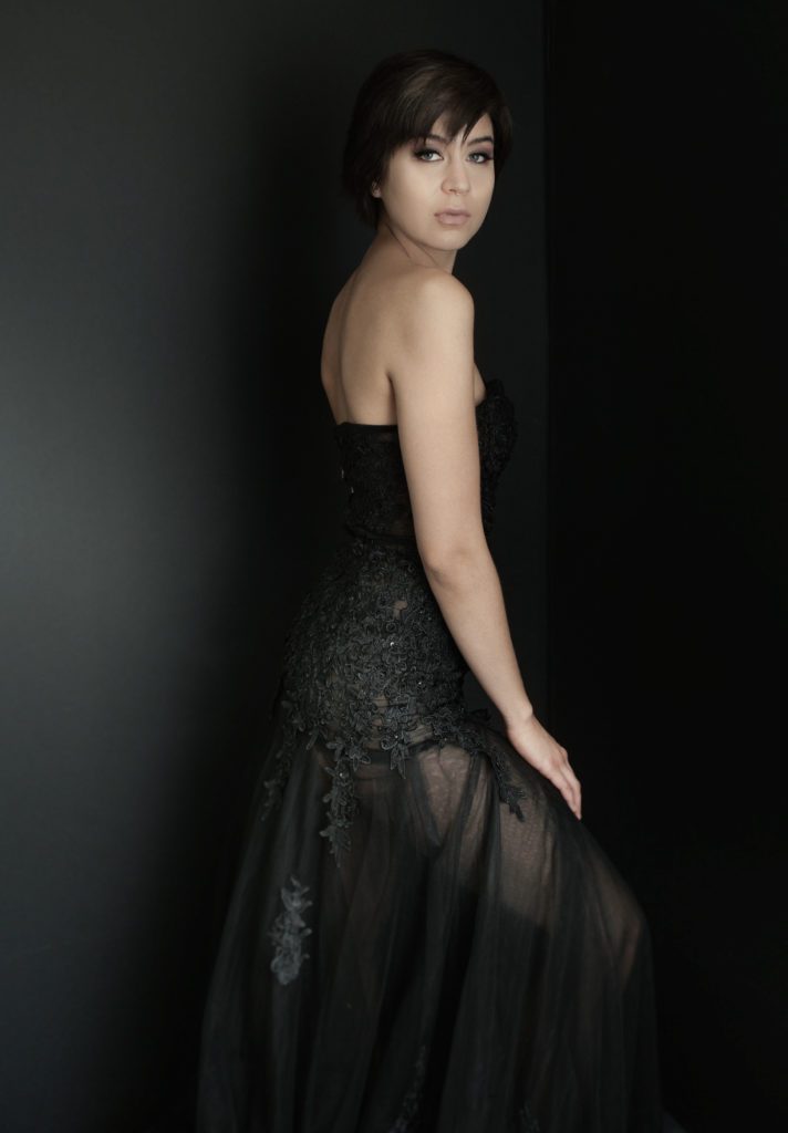 This image was crated at my Pasadena photo studio. It's an image of a young woman in a long black dress. The dress is made of black fabric and tulle and dotted with sequins and black appliques. She is standing half in shadow, but her face is well lit. The dress is shoulder and sleeveless. She is turned to the side and she is looking slightly over her shoulder. Her black hair is cropped into a pixie cut. The background is black.