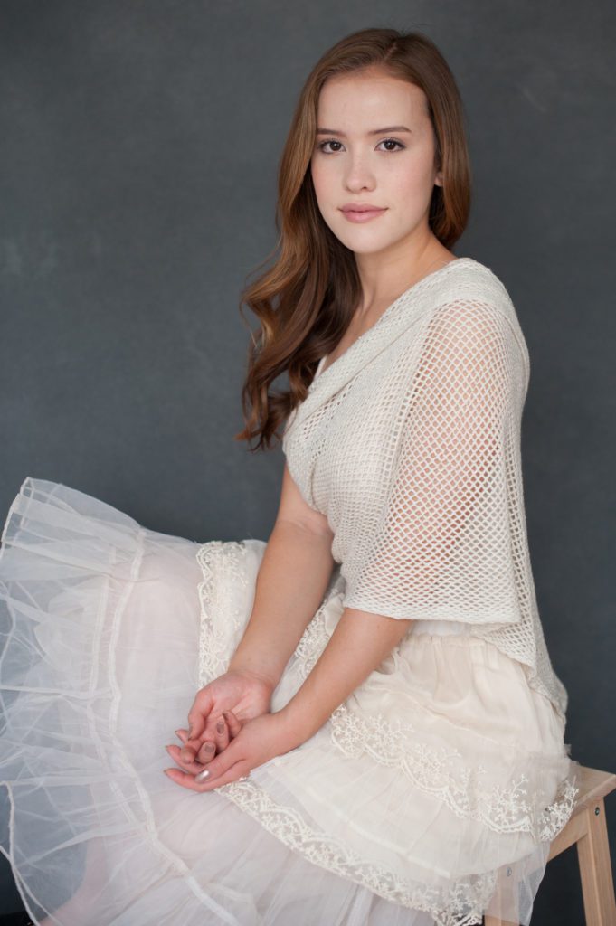 This is one of a series of family photos created in Pasadena, CA in my photo studio. This young teenaged girl is sitting on a stool. She's facing the side. She has on a cream, sheer shawl and long structured, tiered tulle skirt. her hands are resting in her lap. Her long hair is reddish brown. Her skin is pale and her eyebrows are nicely groomed. She has a pleasant expression but is not smiling. When I create family photos, I always take time to create individual portraits as well. Portrait created by Rebecca Little Photogarprhy Pasadena, CA.