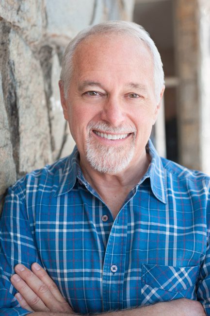 This is a headshot of a man in a bright blue button down shirt. The shirt has a pattern of white and red stripes and looks like professional dress. He is standing outside against a neutral stone wall. He has a white beard, pale skin, and blue eyes. He's smiling an dlooking directly into the camera with an open expression. His arms are crossed. Image created in Pasadena, CA by Rebecca Little Photography.