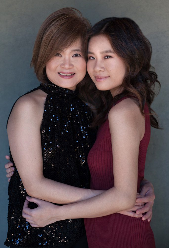 Rebecca Little, photographer for women in Pasadena, CA, created this mother daughter portrait. The woman on the left is wearing a black sequined sleeveless top and her daughter is wearing a sleeveless red ribbed sweater. They are facing each other and their arms are around each other. Their faces are facing the camera and they are smiling.