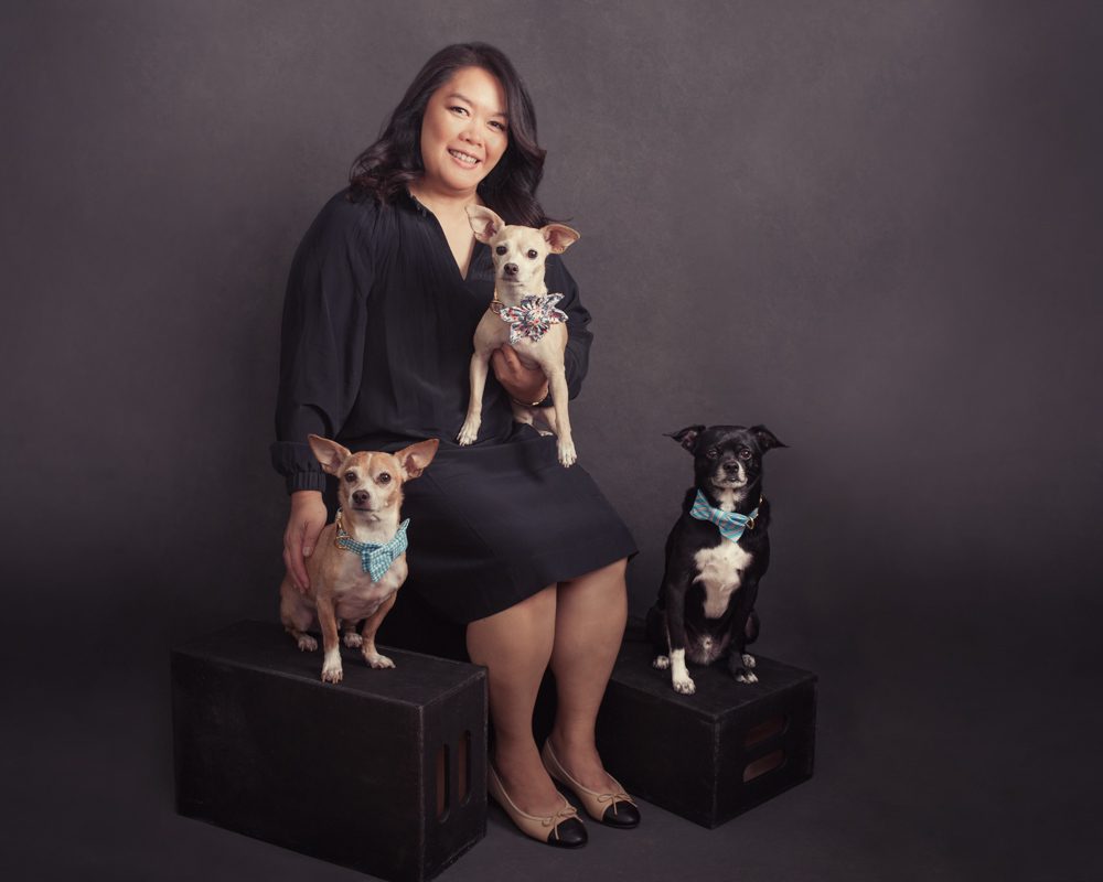 This is a photograph of a woman with her three dogs. I created this pet photograph in Pasadena at my studio. Her dogs are small and she is sitting on a black apple box. One dog is on her lap, the other two are beside her She's wearing a black dress that ends just above her knees and she's facing the camera. She has long black heir and smiling at the camera. This image was created by Rebecca Little Photography in Pasadena, CA.
