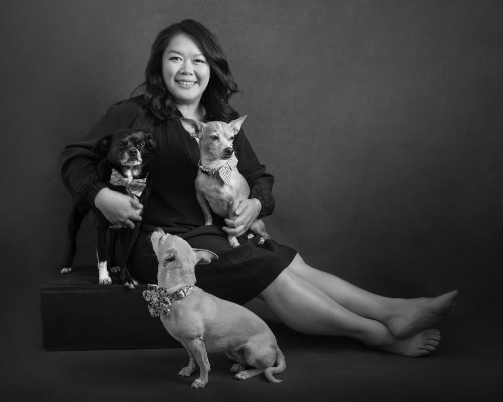 This is a black and white image of a woman with her three dogs. I created this pet photograph in my Pasadena studio. Her dogs are small and she is sitting on a black apple box. One dog is on her lap, the other is beside her, and the third dog is on the floor looking up at her. She's wearing a short black dress and she's facing the side. We can see her bare legs stretched out to the side. Her feet are bare. She has long black heir and smiling at the camera. This image was created by Rebecca Little Photography in Pasadena, CA.