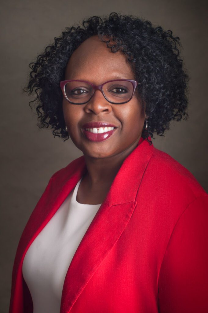 This is a portrait of a Black woman. She has spiral curls that attractively frame her face. She's wearing a red suit jacket. She has maroon glasses and is wearing red lipstick. She has a white blouse under her jacket with a high scoop neck. She is smiling and facing the camera. She looks professional and pretty. This headshot was created by Rebecca Little Photography Pasadena, CA.