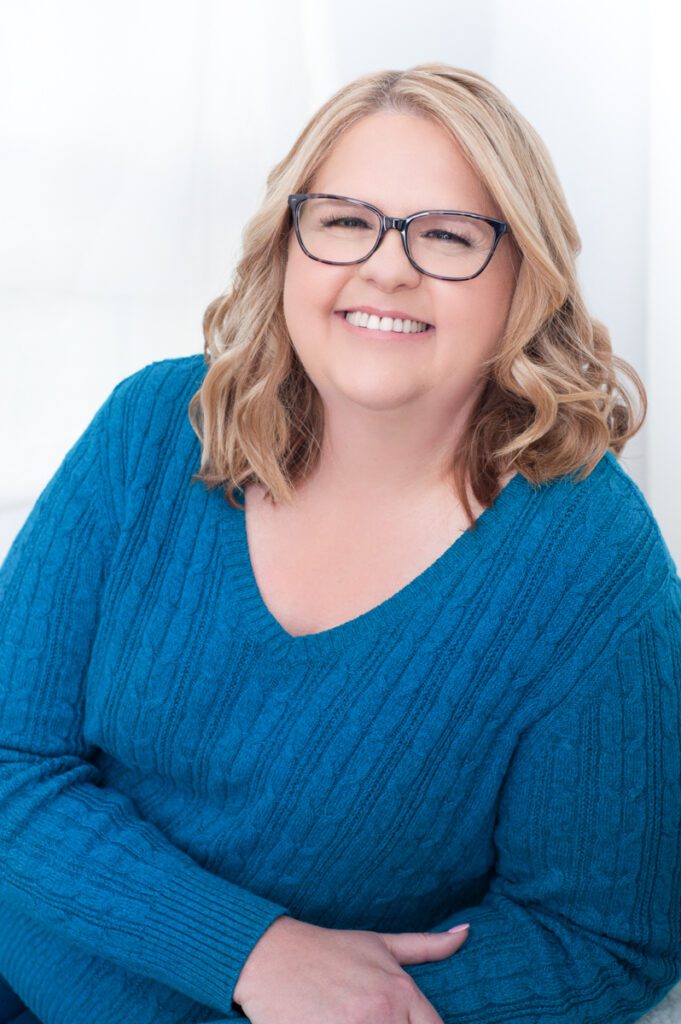Jenni is a business owner in Wrightwood. She's wearing her branding color of bright, happy blue. She has a big smile showing white teeth. She wears dark gray patterned glasses. She comes across as friendly and professional.  Her blond hair reaches past her shoulders. This headshot was created by Rebecca Little Photography Pasadena, CA.