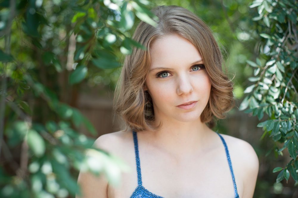 This is an outdoor senior portrait for high school senior Kayla. She is standing outdoors surrounded by greenery. The branches and leaves from the Chinese Elm are framing her face in the most attractive manner. She's wearing a bright blue gown studded with sequins. It has spaghetti straps. He skin is pale and her eyes are blue. She has light brown hair parted at the side and reaching just above her shoulders. The sunlight is soft on her face. She has a winsome expression on her face. Her head is tilted up at a slight angle. Most high schoolers want outdoor senior portraits because they feel more relaxed and natural. I like choosing great locations for outdoor senior portraits because the right setting conveys a certain mood. These soft green leaves portray a gentle mood and softness that compliment her personality. This high school senior portrait was created by Rebecca Little Photography in Pasadena, CA.