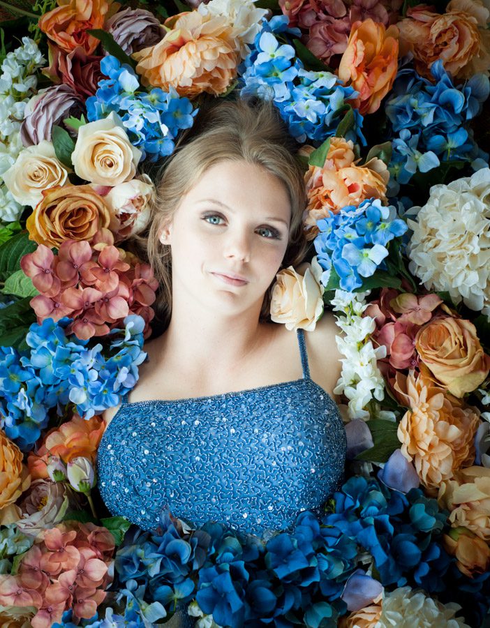A senior portrait for Kayla, the Sierra Madre Rose Float Princess. This young woman, a year out of high school, is lying in a bed of flowers. The flowers are shades of blue, peach, yellow, orange, red, and white. She's wearing a pale blue gown studded with sequins. The dress has spaghetti straps. The flowers are covering he torso up to her bust line and all around her head. Her hair is blond and pulled back behind her. Her eyes are pale blue. She has a slight smile and is gazing at the camera. Image created by Rebecca Little Photography Pasadena, CA.