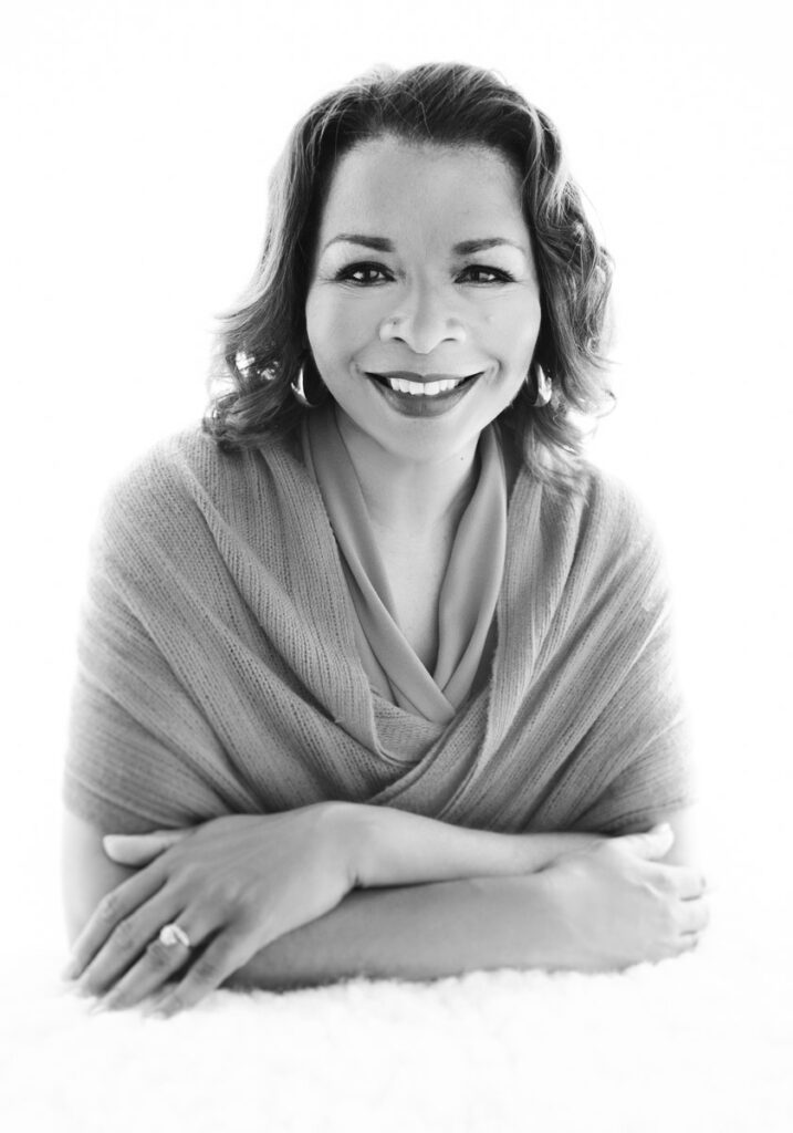 This is a branding image for a Pasadena accountant. It's a black and white image and the client is Black. She has a scarf wrapped around her shoulders and it's tucked into her arms so that it looks like a sweater. Her arms are crossed in front and she's wearing her wedding ring. It sparkles. Her hair reaches to her shoulders and is swept back from her face. She has a direct gaze and smiles at the camera. This headshot was created by Rebecca Little Photography Pasadena, CA.