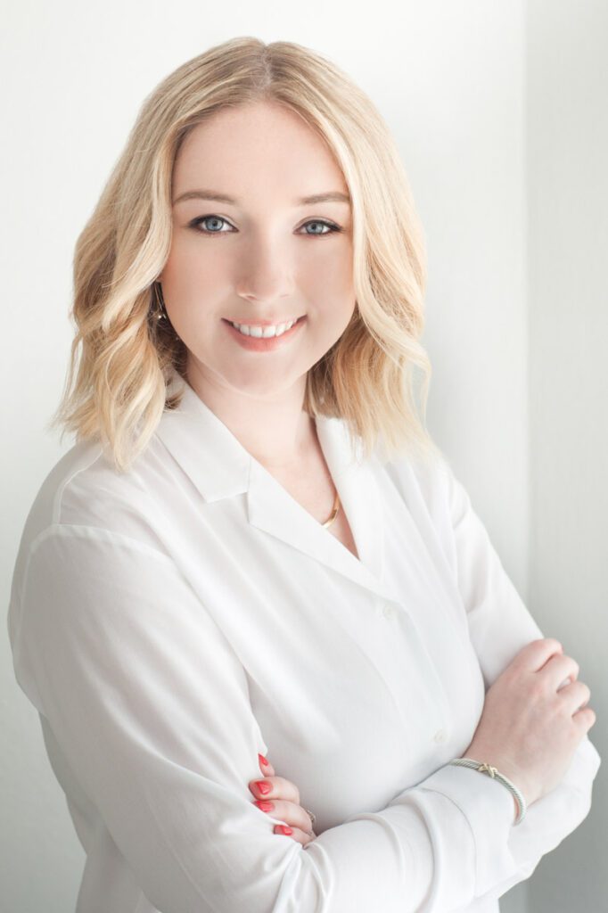 This is an image of a young woman in her 20's and shows how important clothes for headshots are. She has pretty blond hair that just skims her shoulders. It's highlighted in front to frame her face. It's been styled with soft curls. She's wearinf a white blouse with a v-neck and long sleeves. The sun is shing through the window and making the blouse very bright. Her arms are crossed. She has painted her nails a bright red and is wearing a silver bracelet on her right wrist. Her body is turned 45 degrees from the camera and she is looking directly into the camera. Her eyes are blue, her skin is pale and washed smooth with the sunlight. Her eyebrows have been nicely groomed to frame her face and are a dark blond. She's wearing small silver earrings that are barely visible. We talked about what wardrobe to bring for her session because clothes for headshots are important. This white blouse gives her a fresh, modern look and works great with her skin, eyes, and hair color. Headshot created by Rebecca Little Photography Pasadena, CA.