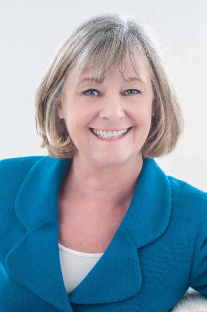 This is an image of Margaret McAustin, former Pasadena city council member. She's wearing a bright turquoise blue textured jacket. The lapels are curved and feminine. There's a white scoop-neck t-shirt underneath. This is a good example of why clothes for headshots are so important. Look how flattering this outfit is on her. She's wearing tiny silver stud earrings. Her hair is short, blond, and curls nicely into her neck. She has a big smile and is looking directly at the camera. The sun is coming from behind her from my big studio window. The blue of her eyes is  set off by the blue jacket. I wanted to create a nice bright headshot that had a happy approachable feel.This image was created by Rebecca Little Photography Pasadena, CA
