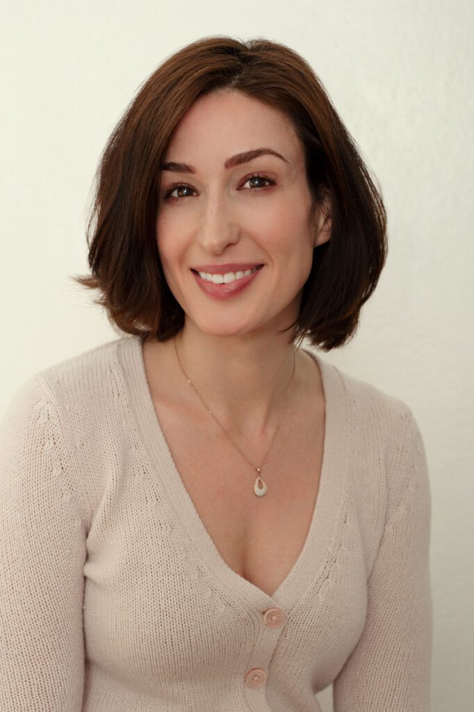 This image is from a series of acting headshots. Meredith is wearing a soft ivory sweater and a gold chain with a simple pendant. Her dark hair is bobbed just above her shoulders. Her eyebrows are nice styled and she's wearing a soft pink and brown eyeshadow. Her skin is smooth and pale. She has on pink-rose lipstick that compliments her coloring. She's smiling at the camera. This headshot was created by Rebecca Little Photography Pasadena, CA.