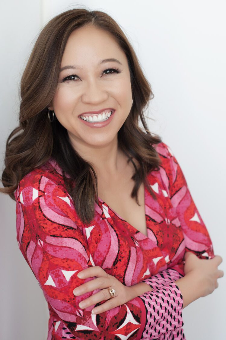 An image of a Vietnamese woman i her forties wearing a bright pink, red, and white dress. it has a v-neck and long sleeves. Her arms are crossed. She's facing the camera with a big smile on her face. Her hair is long and dark brown, almost black. The backdrop is a white wall. This headshot was created by Rebecca Little Photography Pasadena, CA.
