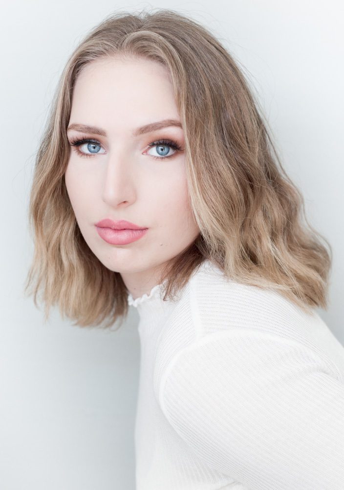 Portraits for teen girl Shelby. Shelby is wearing a white turtleneck. Her hair is blond. Her eyes are bright blue. She is standing against a white wall and the effect is bright and white. Her lips are coral colored and expertly outlined. Her body is turned to the left and her face is turned toward the camera. Her gaze is neutral. Photo created by Rebecca Little Photography in Pasadena, CA.
