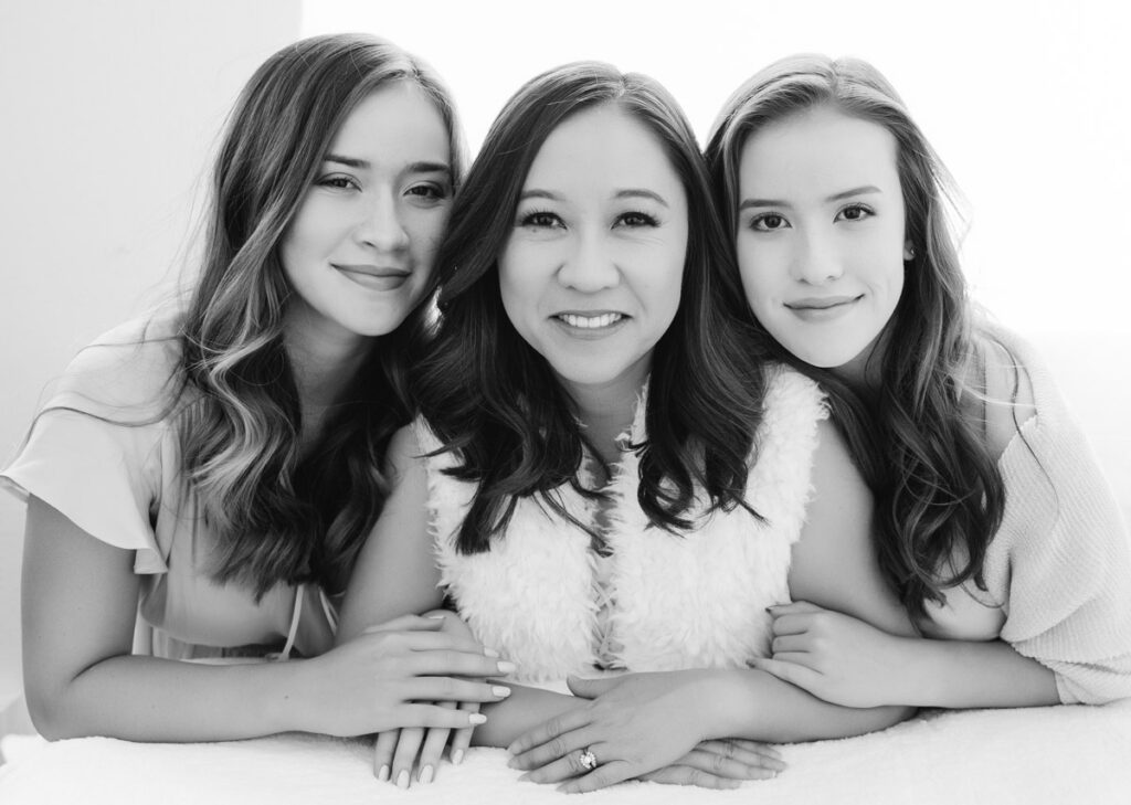 A black and white family photos created in Pasadena CA. This is a black and white image of a mom and her two daughters. They are leaning forward and resting their arms on a table covered in white cloth. The mom is in the middle. They are all dressed in white. The teenaged girls are either side have long brown hair. Mom has hair past her shoulders and it is darker than her daughters'. They are smiling at the camera. The light is streaming through a large window behind them and filling the room with light.  Their skin is soft and smooth. This is a fabulous example of backlighting. Backlighting is gorgeous and flattering and perfect for family photos and mother and daughter portraits. Image created by Rebecca Little Photography Pasadena, CA.
