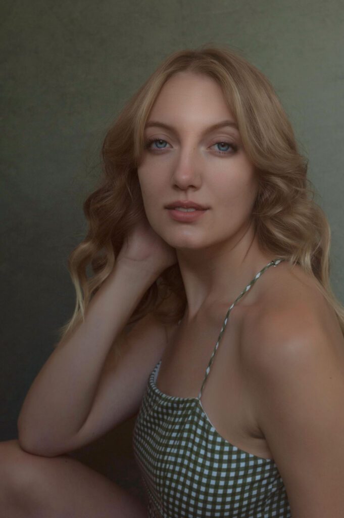 This is a photo of J, a woman in her early 20's. She's wearing a green gingham top with spaghetti straps. She is slim and her blond hair is in soft waves past her shoulders. Her eyes are a striking blue. Her brows are brown and groomed nicely to frame her eyes.  The lighting is coming from above right. The lighting is painterly and diffused. She's looking directly at the camera with just a wee smile. Her expression is pleasant and confident. Her back elbow is on her back leg and her hand is on her neck, partially obscured by her hair. Image created by Rebecca Little Photography in Pasadena, CA.