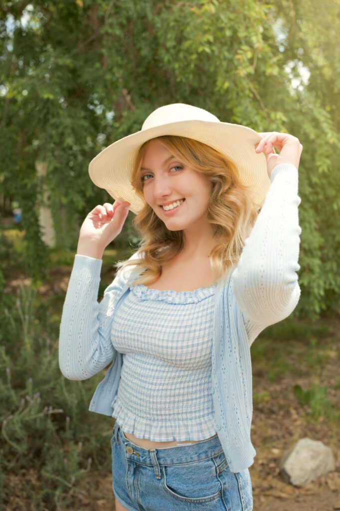 This is an image of J who wanted online dating photos. She standing with the sun behind her and wearing a sporty wide-brimmed ivory-colored hat. She has on blue jean shorts, a smocked light blue gingham top, and a light blue sweater with delicate eyelets.There is a willow tree behind her and the leaves provide a nice backdrop. The sun is coming from the right and is casting a soft golden light on her shoulders and hat. She's smiling at the camera. The photo shows her from the bottom of her shorts to above her head. She's holding the brim of her hat with both hands and smiles flirtatiously at the camera. Her hair is golden blond and styled in soft waves. This is the perfect example of a photo for an online dating site because it shows her body and face clearly and she looks friendly and approachable. Image created by Rebecca Little Photography in Pasadena, CA.