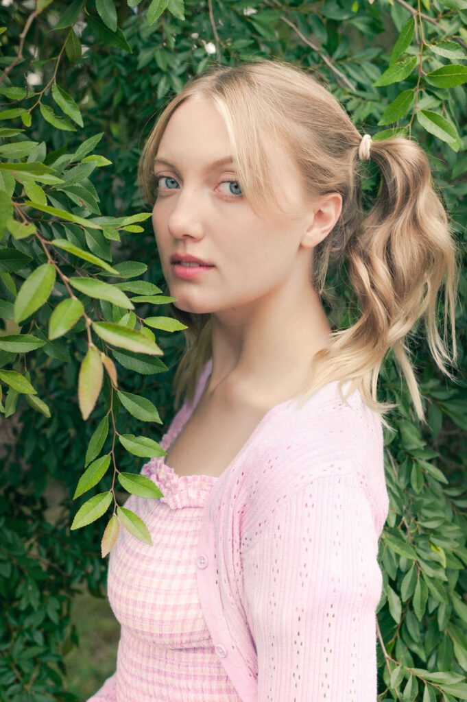 This is an online dating photo created for J, a young woman in her early 20's. She standing among the green leaves of a willow tree. Her blond hair is in two ponytails. The ponytails are on either side and up high and she has bangs sweeping across her eye. She is facing to the left and her face is turned three-quarters to the left. Her blue eyes are looking straight into the camera. Her skin is pale. She's wearing a light pink smocked top and a light pink open-front sweater. Her lip color is bright pink. Image created by Rebecca Little Photography in Pasadena, CA.