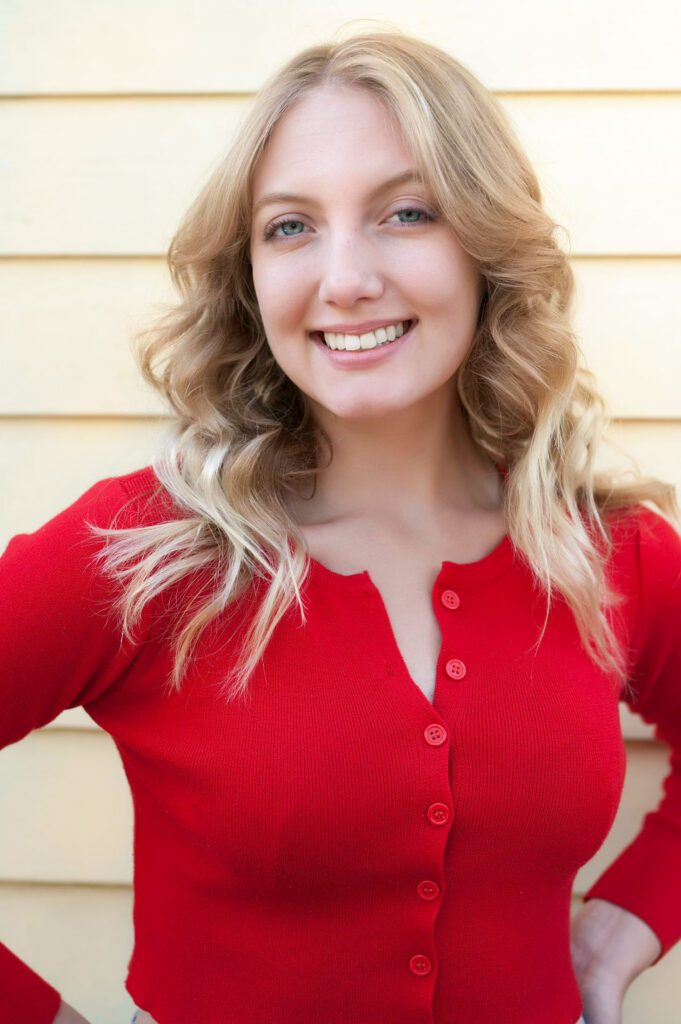 This is an images of a young woman wearing a red sweater. The sweater has long sleeves and is buttoned up. The woman has long blond hair and is smiling at the camera. She is standing in front of a yellow wall and smiling at the camera. Here eyes are blue and her skin is pale. This is an example of an outdoor informal headshot that is used for a dating site. This is an example outfits to wear for dating photos as it is a happy confident color and looks good with her hair and skin. Image created in Pasadena, CA by Rebecca Little Photography.