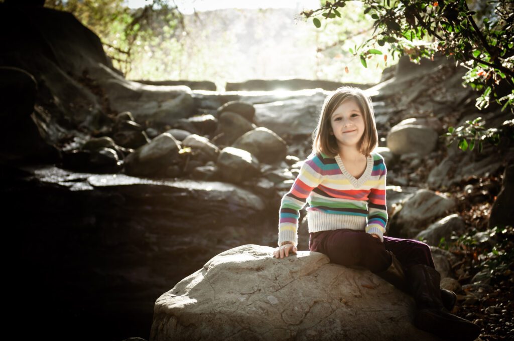 One of a series of family photos created at Descanso Gardens in La Canada, CA. his little girls is wearing a brightly striped sweater. She's sitting with a waterfall behind her. The sun is behind her and shining on her hair and shoulders. She's sitting on a large boulder and smiling at the camera. Photo created by Rebecca Little Photography Pasadena, CA.