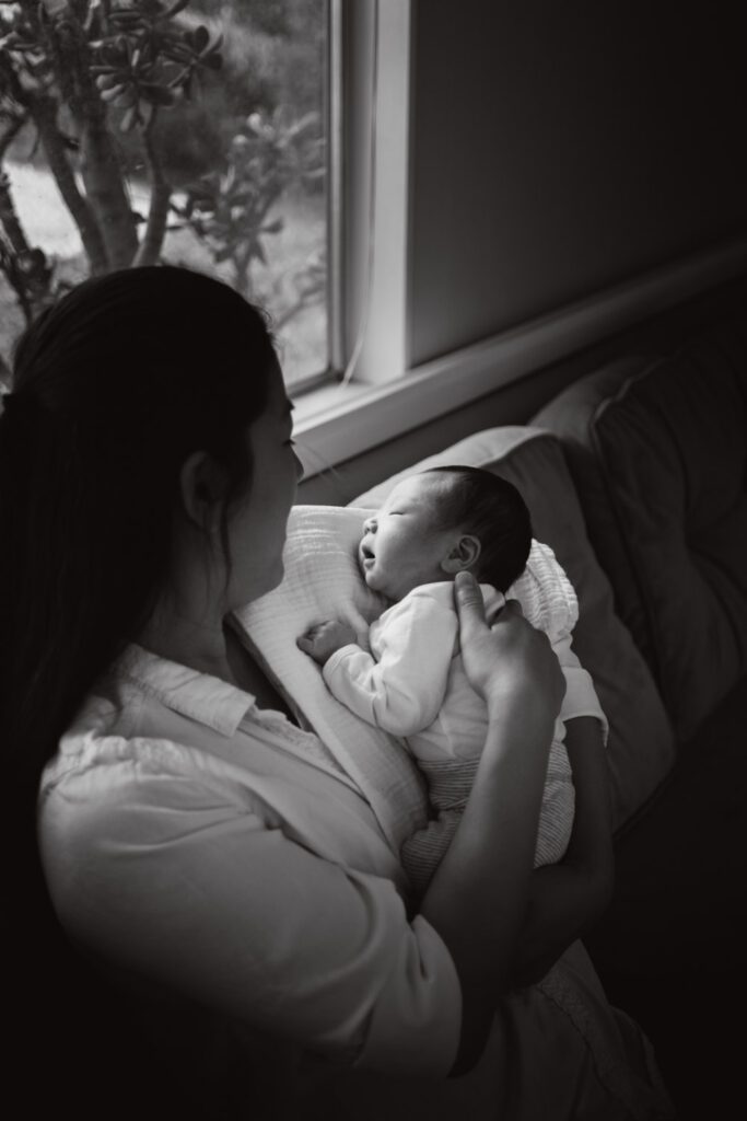 A black and white portrait taken during a newborn lifestyle session in Sierra Madre, CA. The mom and baby are sitting on a sofa in front of a window. The baby is asleeping on mom's shoulder. The sunlight is illuminating his face.
