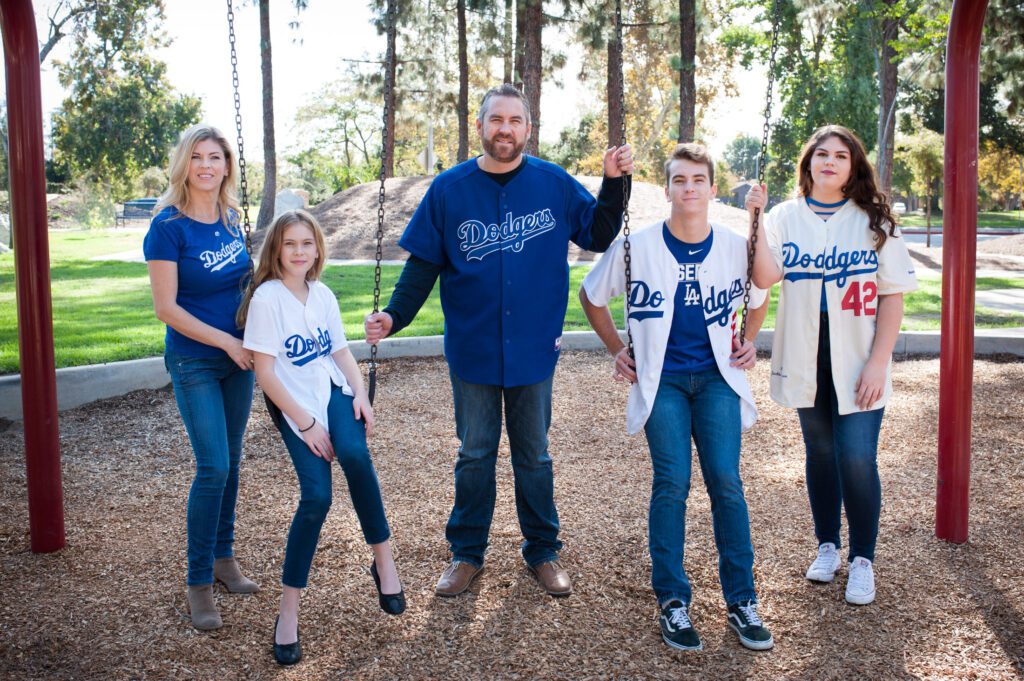 A family of five posing at an outdoor park in Pasadena, CA. They are wearing Los Angeles Dodger gear and are sitting an dstanding at a swingset. The Dodger outfits are blue and white. They are all wearing dark blue jeans. Dad is standing in the middle and mom is on the outside left. Their youngest daughter is betwen them and she's sitting on a swing. Their two other children, a teenaed boy and girl, are on the other end. She is standing on the end and he is sitting on the swing. They're looking at the camera and smiling. Behind them is a small dill, grass, and trees. This family photo was created in Pasadena by Rebecca Little Photography.