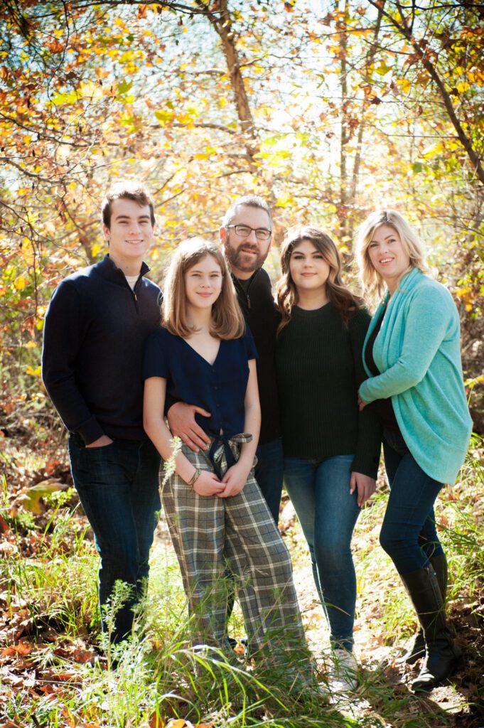 This family of five is at Eaton Canyon in Pasadena posing for family photos. This mom, dad, and three school-aged children are grouped close together. Their outfits are coordinated with black, navy blue, and a teal sweater. In the background are tall trees and branches with green, gold, yellow and brown leaves. Image taken by Rebecca Little Photography Pasadena, CA.