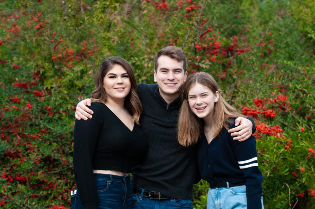 This is an images of three school-aged children posing for a family photo in a Pasadena park. They're coordinated with black and dark blue sweaters and jeans. The brother, the oldest and tallest, is standing in the middle and has his arms around his two sisters. There is a large green bush with red bracts behind them.