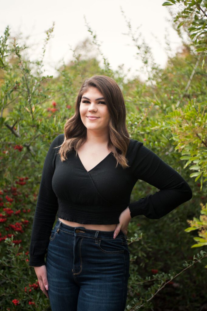 Look at this beautiful outdoor location for family photos! A teenaged girl is standing among green tree branches and red bracts. The leaves surround her and frame her. She's wearing blue jeans and a black sweater crop top. One hand is on her hip/ He blonde-brown hair reaches past her shoulders. The sweater has a crossover V-neck and long sleeves. She's smiling at the camera. Image created by Rebecca Little Photography Pasadena CA.