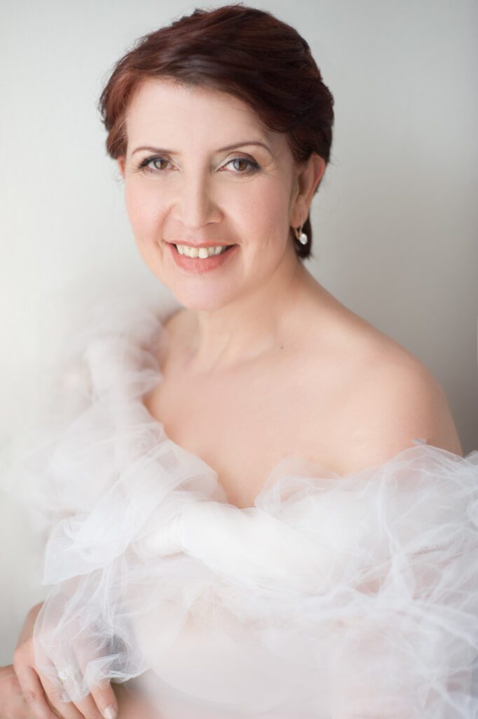 A portrait of an older woman. She's standing to the side of a window and the sun is shining through. She's wearing white tulle wrapped and twisted around her shoulders. One shoulder is bare. Her skin is pale. The light is bright and flattering on her skin. Her eyes are brown and she is smiling at the camera. Her brown-red hair is cropped short. She's wearing delicate earrings with a white stone. This is a good example of flat lighting that's coming from the side. Image created by Rebecca Little Photography Pasadena CA.