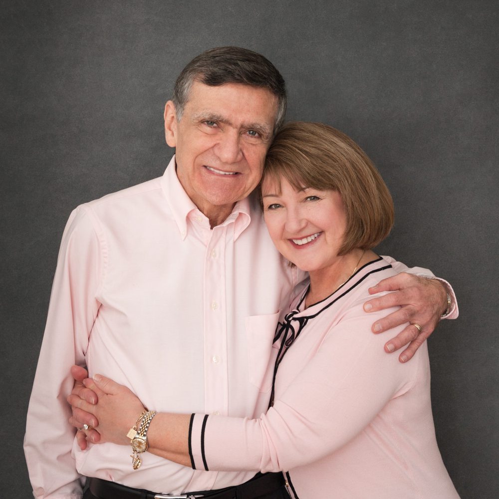 This is a portrait of a husband and wife at my Pasadena studio. He is wearing a light pink buttoned down shirt and she is wearing a matching light pink sweater with black trim. She is standing sideways with he arms around his waist. She has a gold watch and bracelet. They look elegant. They came in for family photos with their three adult daughters, their husbands, and their grandchildren. Image created by Rebecca Little Photography Pasadena, CA.