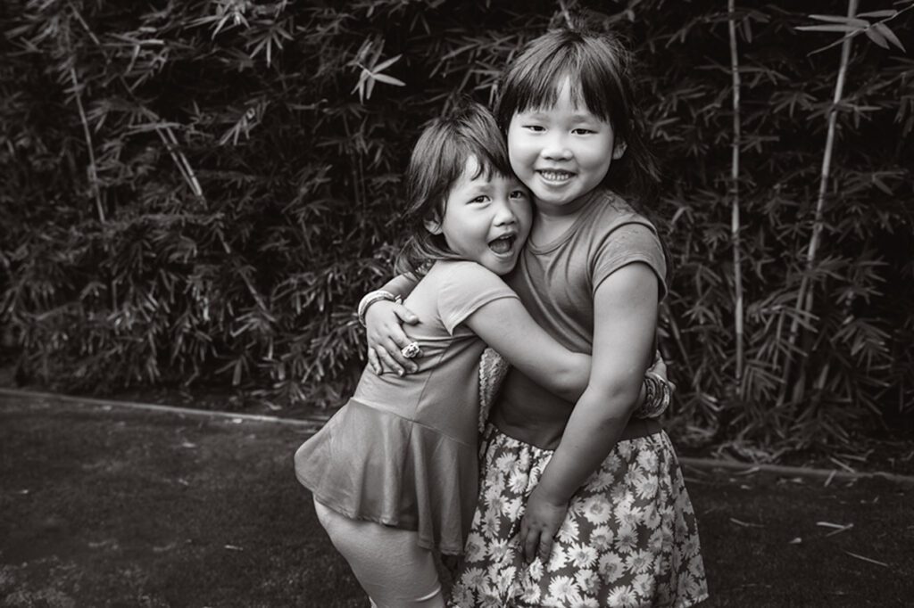A black and white family photo of two young girls at the San Marino backyard of their family. The two girls are hugging each other. There is bamboo behind them and they're standing on the grass. Image created by Rebecca Little Photography Pasadena, CA.