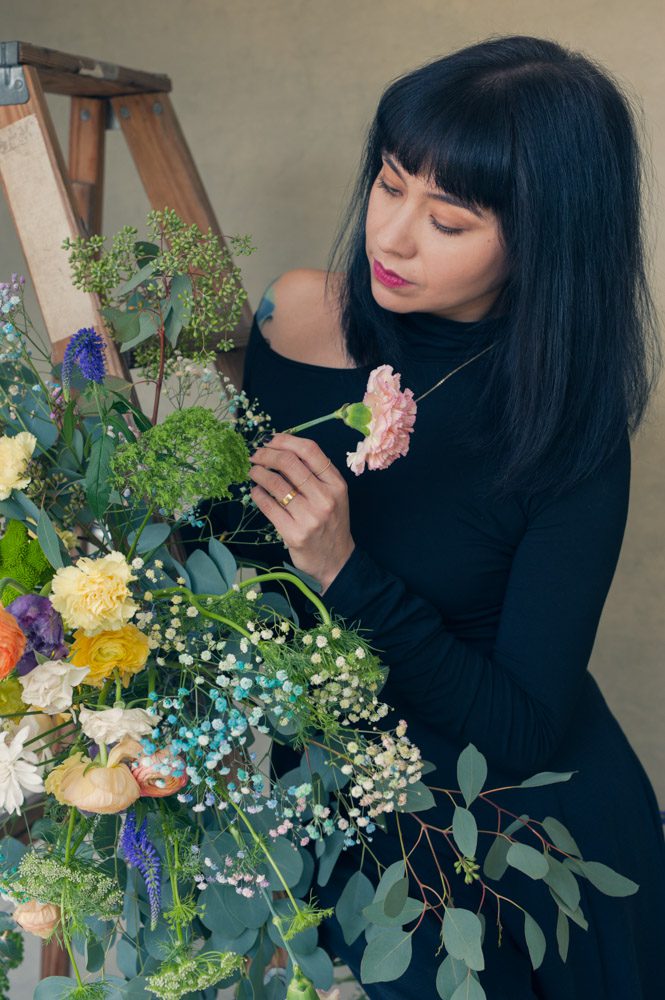 Branding photo for floral designer Bri orozco of Blowouts & Blooms. It's so fun to work with artistic people and visualize and create branding photos that capture their spirit of their business. This florist wanted to display her flowers in an attistic way and we came up with two concpets. In this image, Bri is wearing a black turtleneck and black pants. She has thick black hair in a smooth bob that hits her shoulders. We've arranged flowers and greenery around a wooden ladder. She is standing beside the ladder and placing a single flower into the arrangement. Branding image created by Rebecca Little Photography at her Pasadena studio.