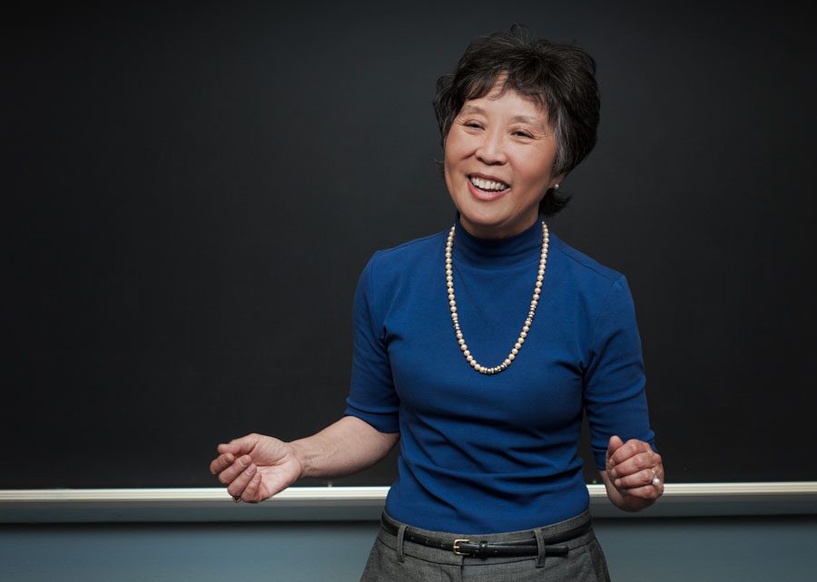 This branding image features Susan, a Japanese lecturer who teaches at USC. She is standing in front of a blackboard and speaking to her students (unseen). She's wearing a deep blue turtleneck sweater with sleeves pushed up to her elbows. She has a long string of pears around her neck. Her hair and short and black. She is gesturing with hands outstretched and a smile on her face. She teaches her students about the Japanese internment during World War II. Branding image created by Rebecca Little Photography at USC.