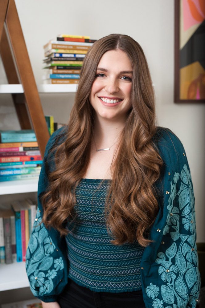 Here is a photo of a young therapist in her office. She has long, thick brown hair with soft waves. She's wearing a deep blue blouse and a gold necklace. She's smiling at the camera. There is a bookshelf behind her filled with colorful books laying horizontally. She is brightly lit and looks relaxed and happy. This Pasadena therapist recently moved to the area and wanted to update her website with new headshots. It's nice to have professional photos done at your business location so potential clients can see where they will be meeting you. This office looks bright, with white walls and colorful books and a painting on the wall. There wasn't enough light from the window so I used my flash to illuminate her face and background.
