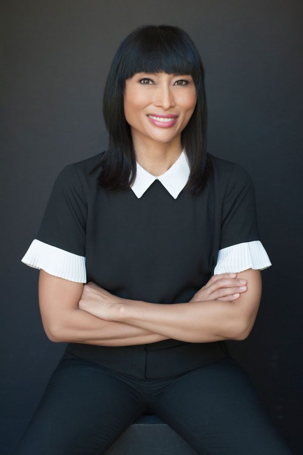 This is a professional image of a woman wearing long black pants and a black blouse with a white pointed collar and white pleated trim on short sleeves. She's sitting on a black apple box and her arms are crossed. She's smiling at the camera. Her straight black hair rests on her shoulders and she has bangs. Her eyes are dark and she has pale skin. The effect is sophisticated, confident, and elegant. This professional updated headshot will be used for a job search. It's always important to update your headshot when searching for a new job or when you're switching careers. You want to look like yourself and you want to feel good about your new portraits. I chose a black background for her image and used filtered sunlight to illuminate her. Image created by Rebecca Little Photography in Pasadena, CA.
